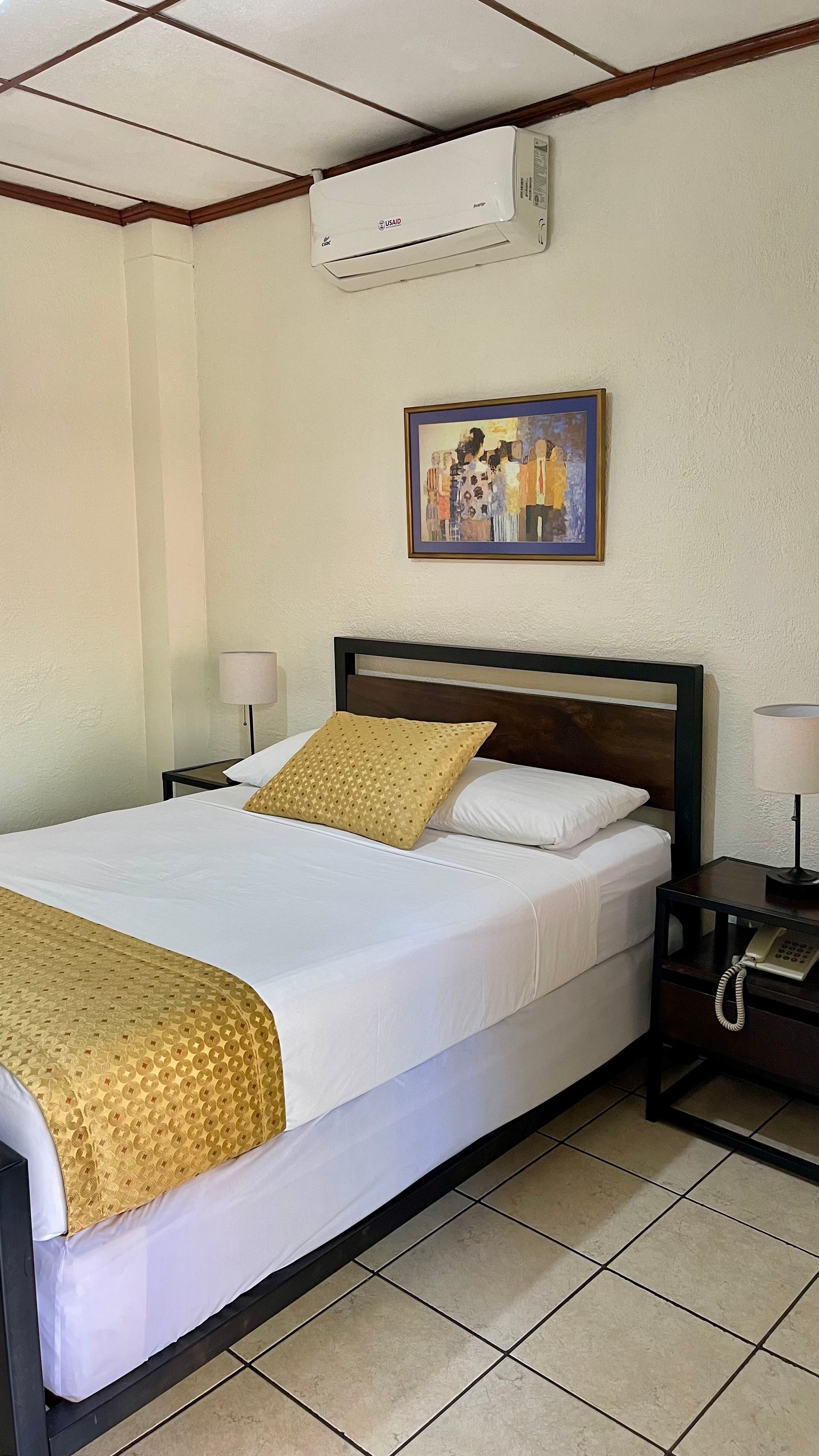 A bedroom featuring a bed with white linens and a gold runner, flanked by two nightstands, a wall painting, and AC unit.