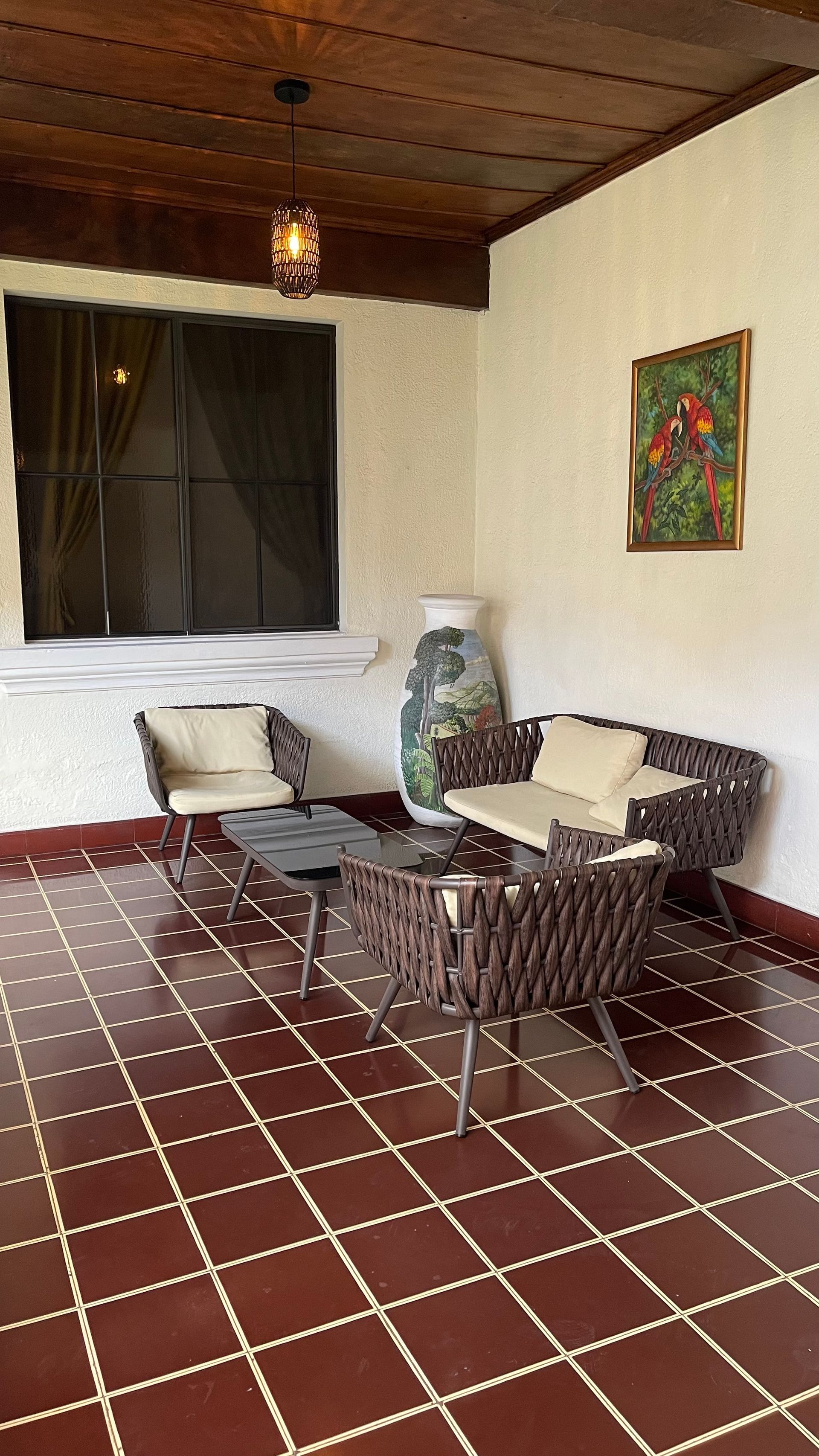 Patio area with dark woven furniture, light cushions, a small glass table, a decorative vase, and brown tile flooring.