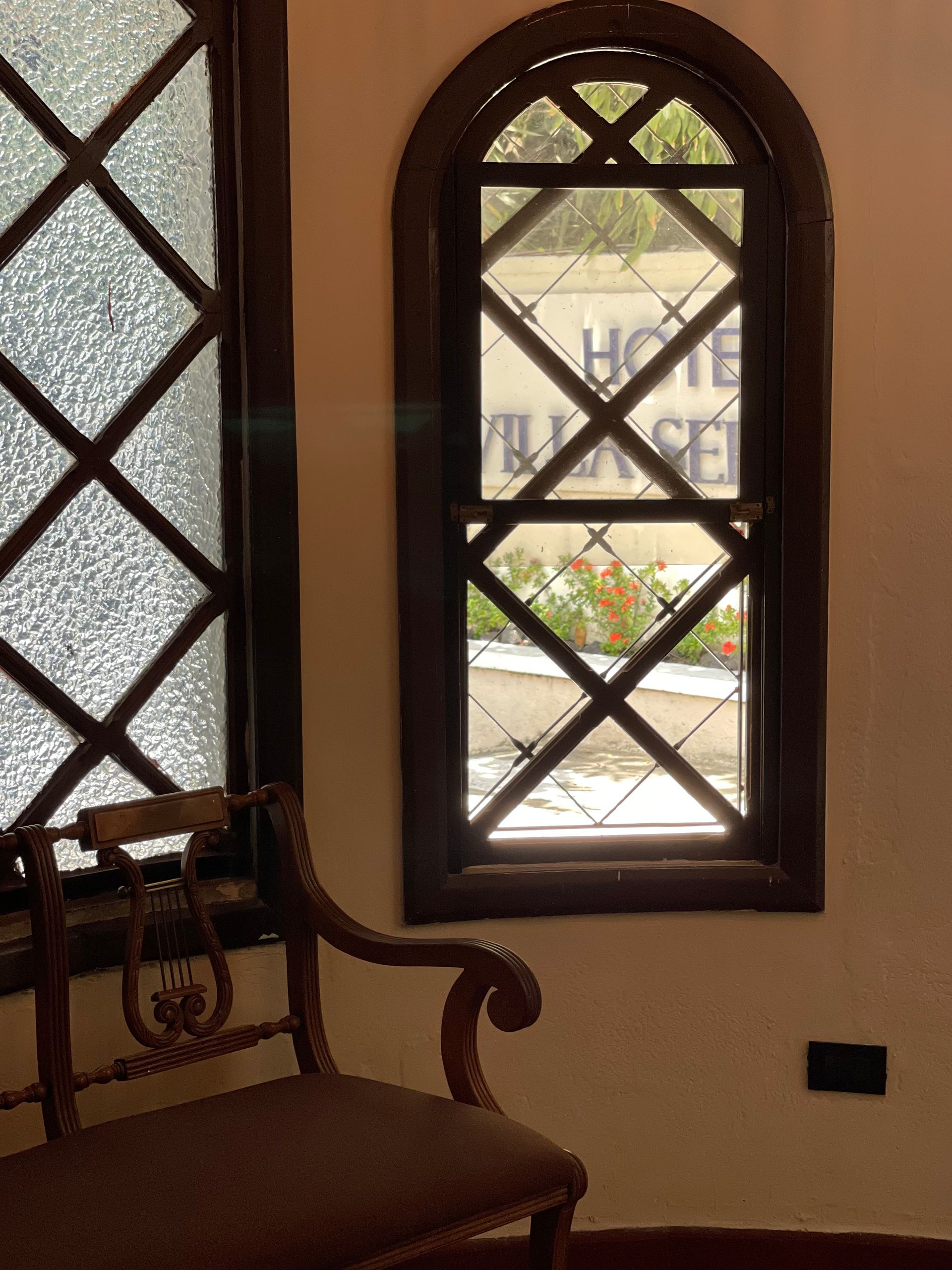 A wooden bench sits next to a window with a diamond-patterned grid, looking out toward a sign for Hotel Villa Ser.