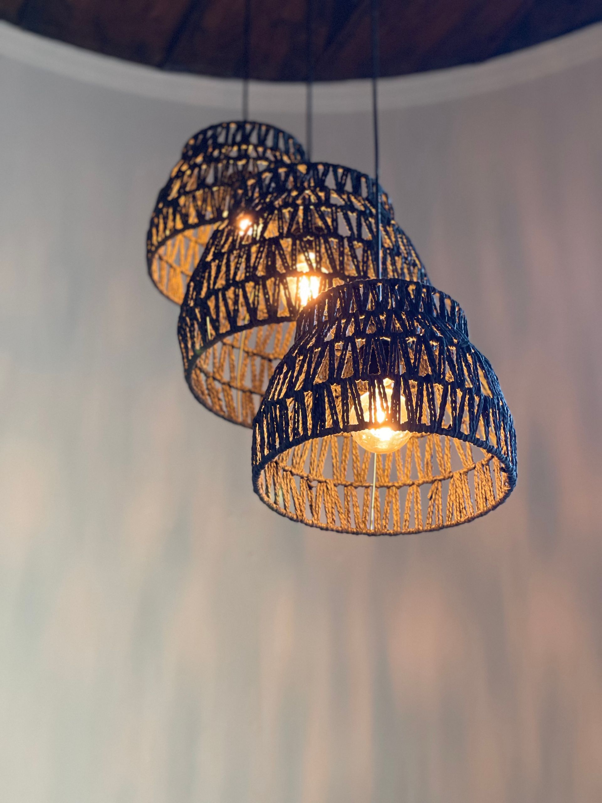 Three layered, woven pendant lights with glowing bulbs suspended against a curved, cream-colored wall.