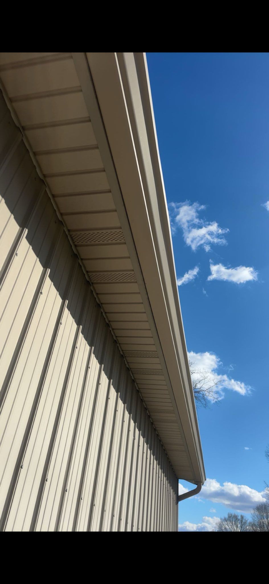 A view looking up at the beige vinyl siding and soffit of a house exterior against a bright blue sky with light clouds.