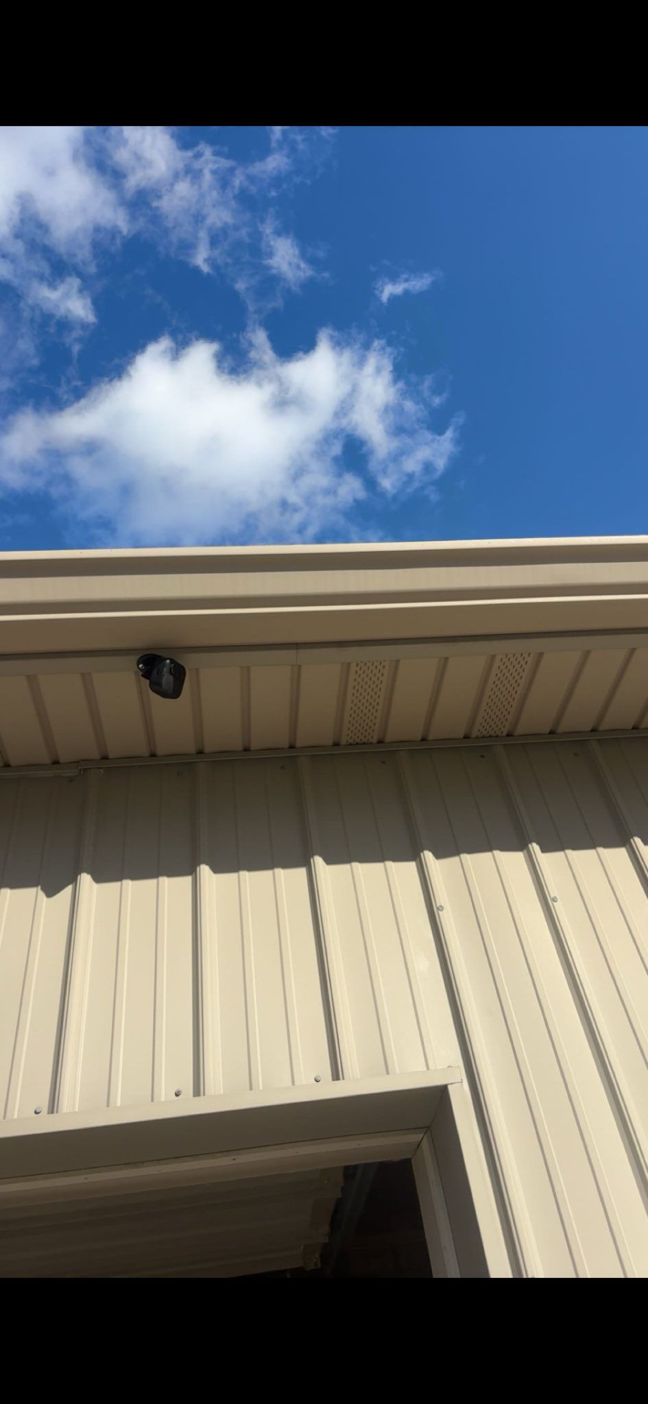 A small dark object, possibly a wasp nest, hangs under the beige metal eaves of a building against a bright blue sky.