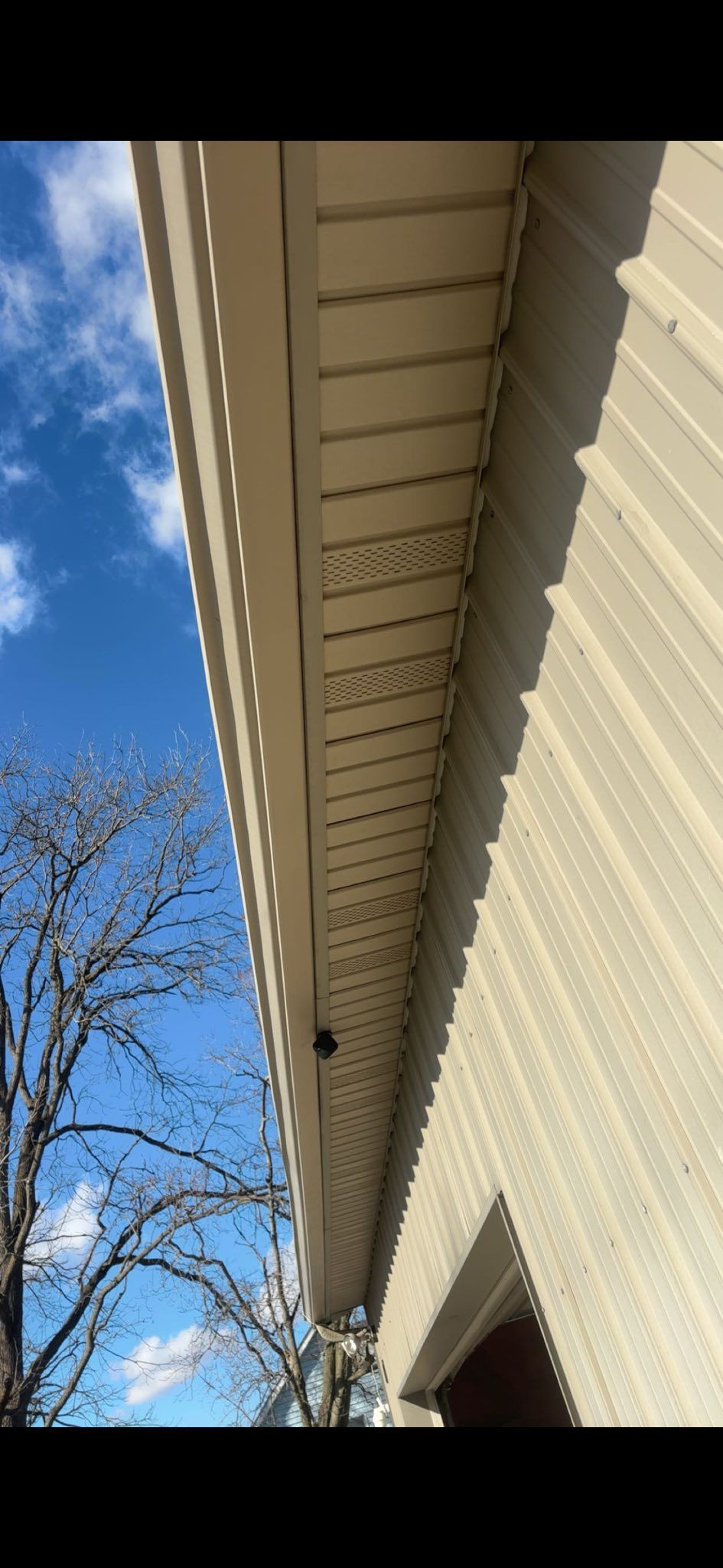A low-angle view of a house eave with beige vinyl soffit, a dark shadow area, and a blue sky with trees in the background.
