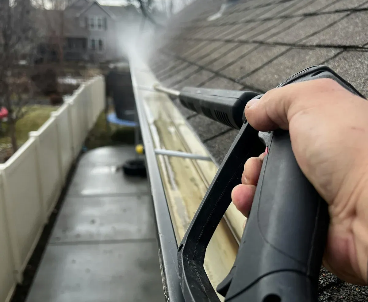 Hand holding a pressure washer cleaning a sloped roof gutter beside a house