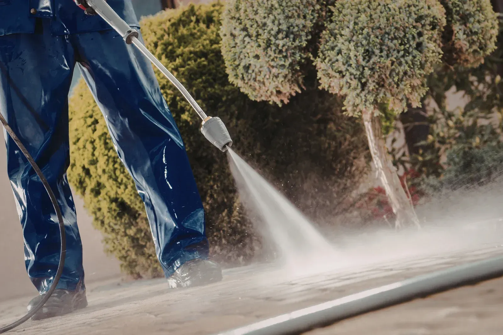 A person in blue protective gear uses a pressure washer to clean a concrete surface near bushes.
