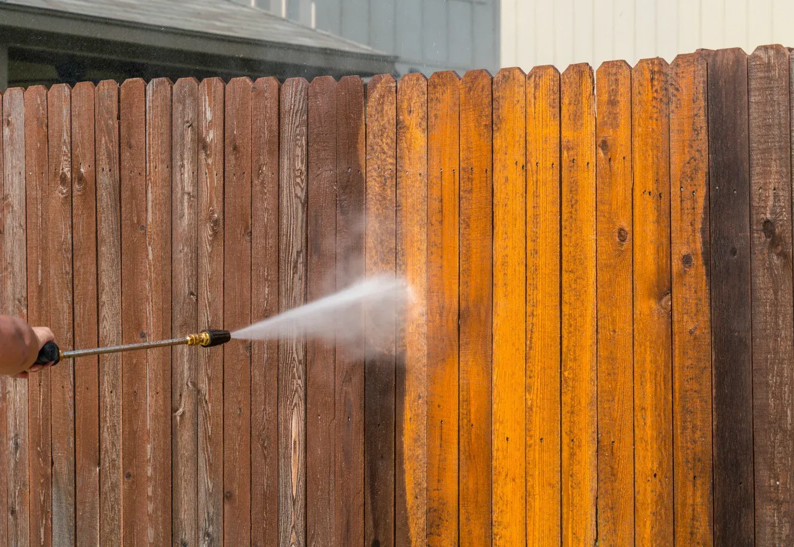 A person uses a pressure washer to clean a weathered wooden fence, revealing bright, clean wood beneath the grime.