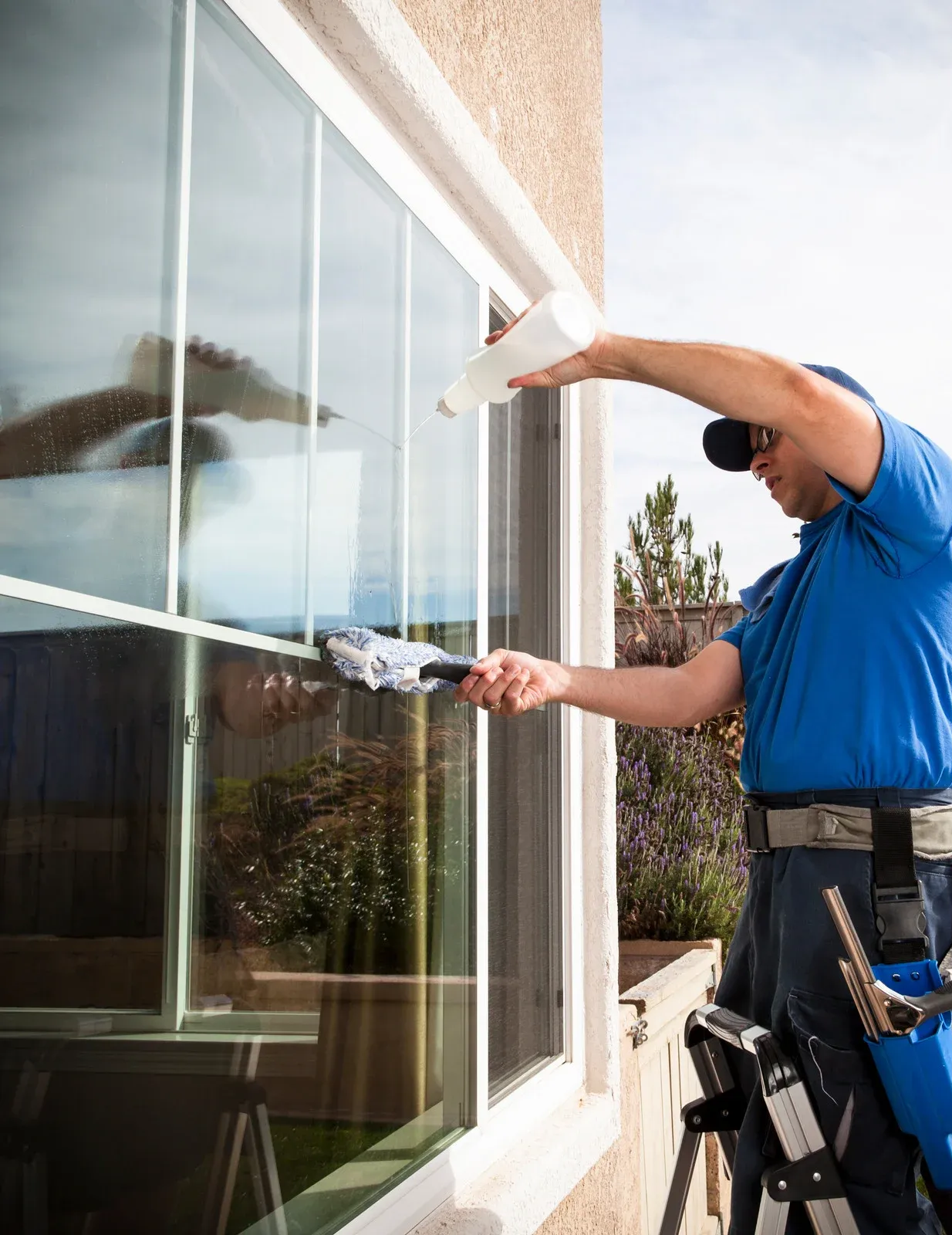 A person wearing a blue shirt cleaning a large exterior window with a cloth and a spray bottle.