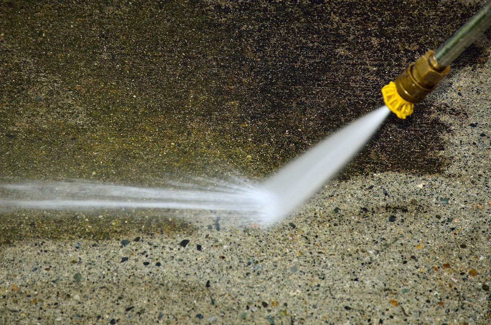 A high-pressure water nozzle cleaning a dark, dirty concrete surface, revealing a light, clean patch beneath.