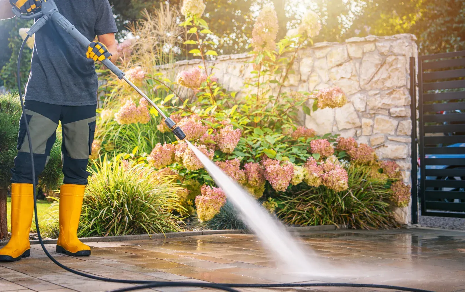A person wearing yellow rubber boots pressure-washes a stone patio in front of a garden with flowering shrubs.