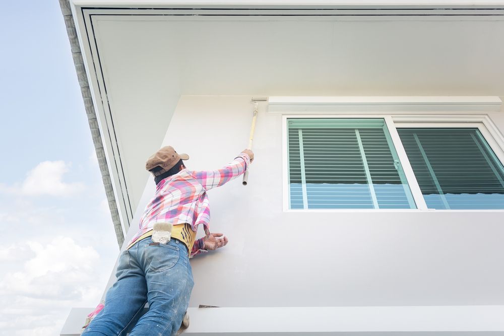 A person wearing a plaid shirt and cap uses a paint roller to paint the exterior wall of a white house near a window.