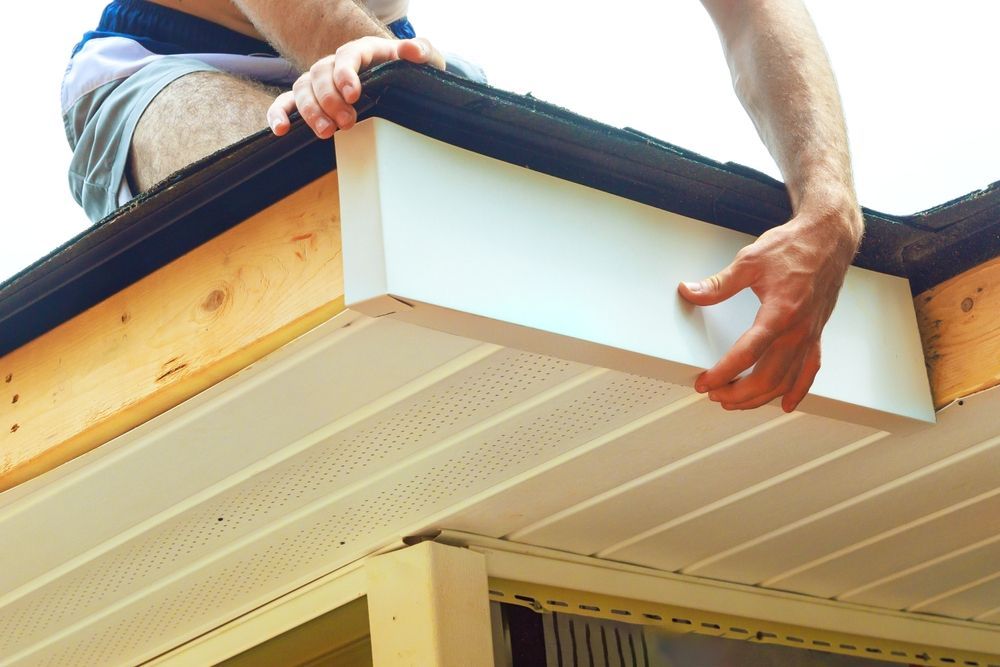 A person installs a white fascia board onto the wooden edge of a roof, next to a vented soffit.