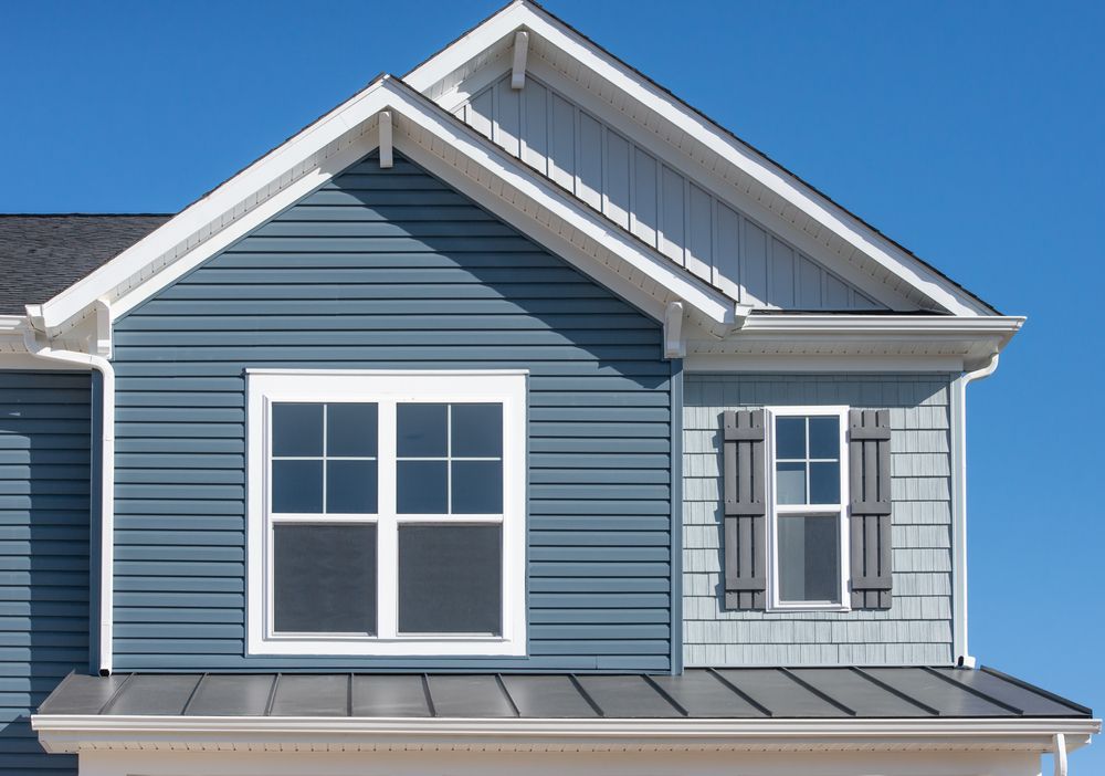 The front upper exterior of a house with blue horizontal siding, white trim, a gabled roof, and a metal porch roof.