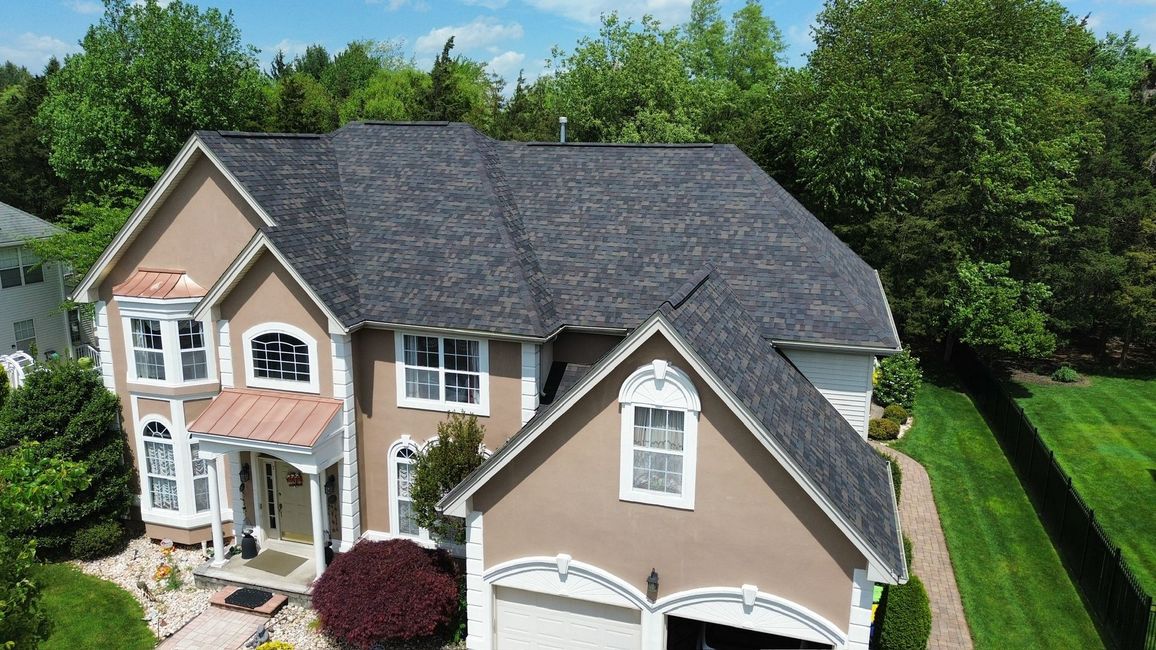 Aerial view of a two-story beige house with a dark shingled roof, arched windows, and green trees in the background.