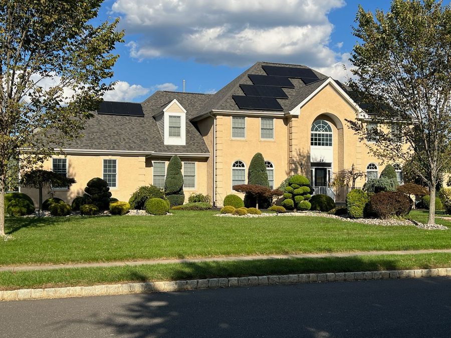 A two-story tan brick suburban house with a gray shingled roof, solar panels, and a neatly landscaped front yard.