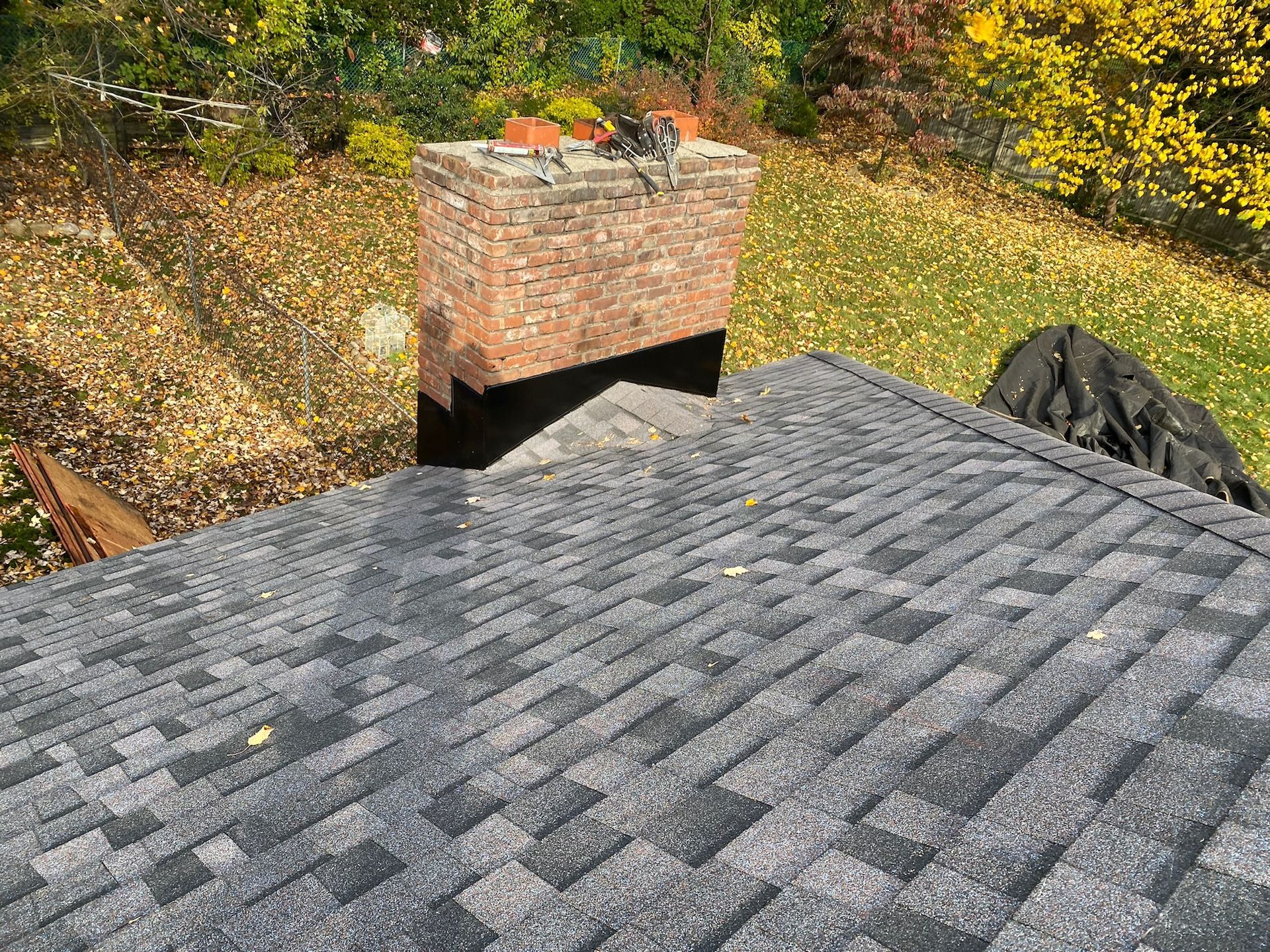 A brick chimney rises from a gray shingled roof, surrounded by a yard with autumn leaves and trees.