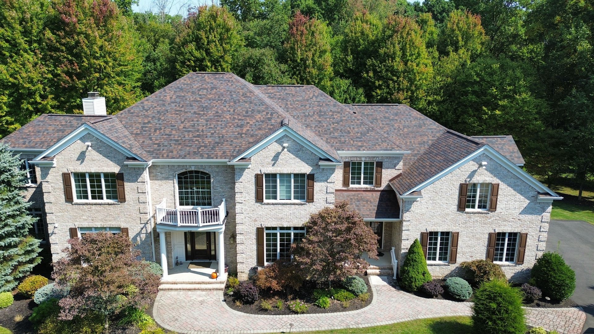 A large, two-story stone house with a complex shingled roof, set against a backdrop of lush green trees.