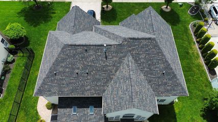 An aerial view of a gray shingled roof on a house, surrounded by a green lawn and landscaping.