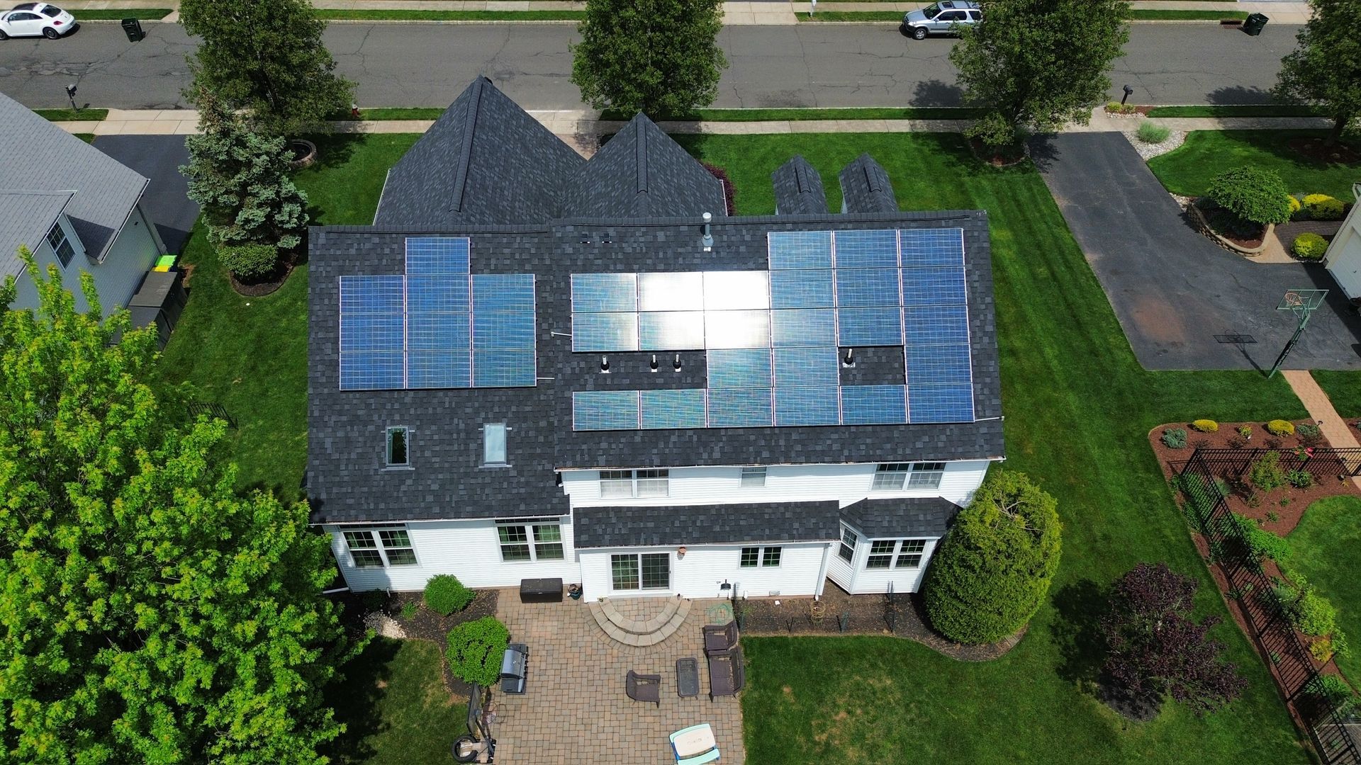 An aerial view of a white residential house with solar panels installed on its dark shingled roof, surrounded by a lawn.