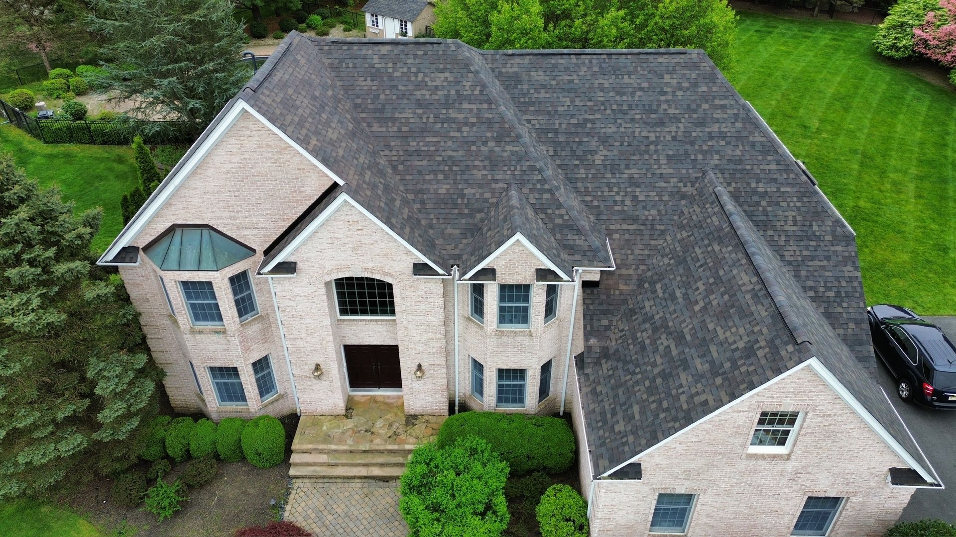 High-angle aerial view of a two-story brick house with a dark asphalt shingle roof, surrounded by a lawn and trees.