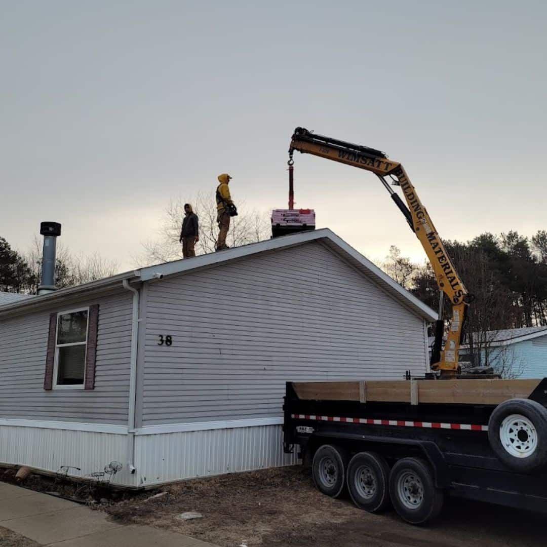 A trailer with a crane on the back is parked in front of a mobile home.