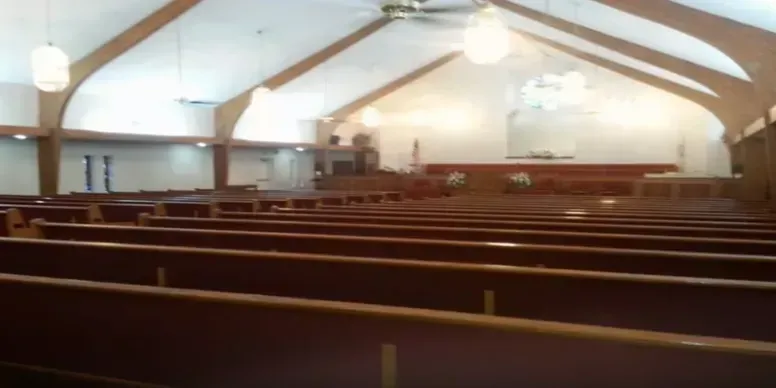 Interior of a church with rows of wooden pews. The ceiling has exposed beams, and a light hangs down.