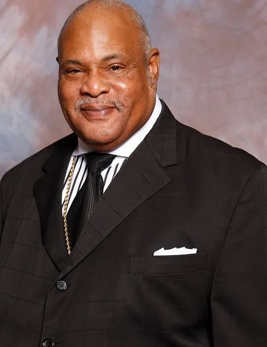 Man in black suit and tie, smiling, indoors.
