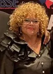 Woman with curly blonde hair and glasses wearing a ruffled black top.