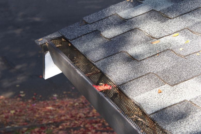 Close-up of a roof gutter with a mesh screen, filled with leaves, next to a gray shingle roof.