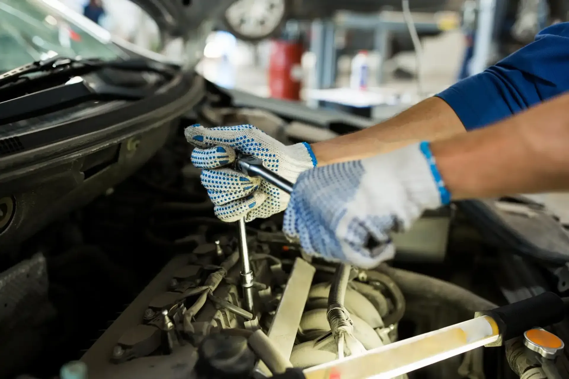 A mechanic wearing work gloves uses a socket wrench to repair a car engine in an auto shop.