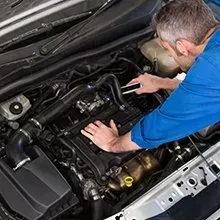 A person in a blue shirt inspects the engine of a car while holding a small flashlight.