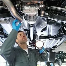 A technician in a grey uniform and blue gloves inspects the underside of a car with a flashlight in a garage.