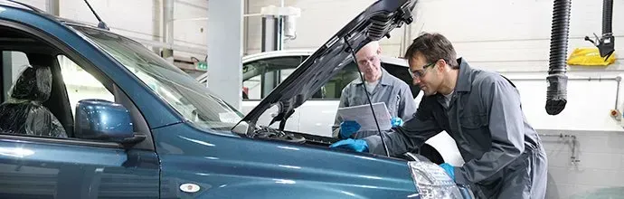 Two mechanics in gray uniforms and blue gloves inspect an open-hooded dark blue car in an auto repair shop.