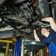 A mechanic in blue overalls works under a car raised on a lift, positioned to catch fluid in a black oil drain pan.