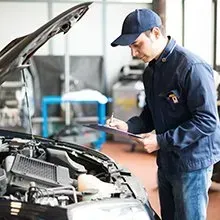 A technician in a blue uniform writes on a clipboard while inspecting the open engine bay of a car in a workshop.
