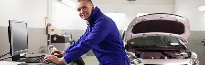 A technician in a blue uniform smiling while typing on a computer in a garage next to a car with its hood open.