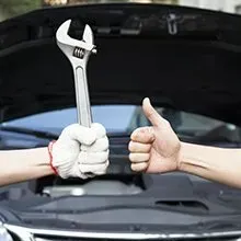 Two hands in front of an open car hood: one wearing a white glove holding a wrench, the other giving a thumbs-up.