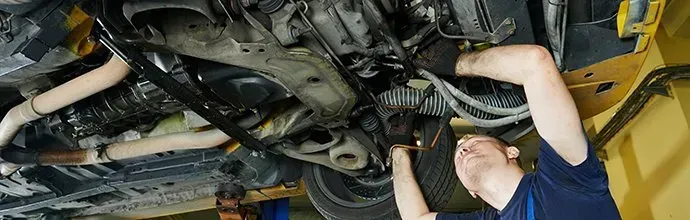 A mechanic in a blue shirt works on the underside of a car raised on a lift in a workshop.