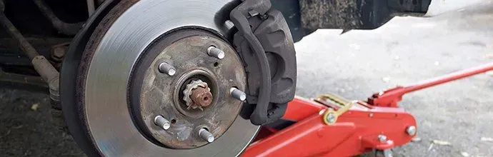 A car wheel hub and brake rotor with a brake caliper attached, elevated by a red hydraulic floor jack.