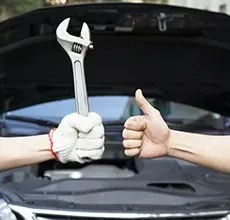 Two hands in front of an open car hood: one wearing a work glove holding a wrench, the other giving a thumbs-up.