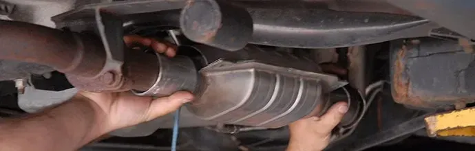 Hands hold a vehicle's catalytic converter in place while connecting it to the exhaust pipe underneath a car.
