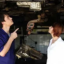 A mechanic in a blue uniform points toward the undercarriage of a car while explaining repairs to a customer.