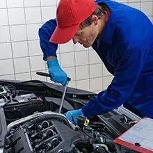 Mechanic in a blue uniform and red cap uses a wrench to work on a car engine in a tiled garage.