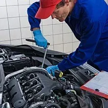 A mechanic in a blue uniform and red cap uses a wrench to work on a car engine in a tiled garage.