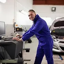 A mechanic in a blue jumpsuit smiling while working at a computer terminal in an auto repair shop with a car nearby.