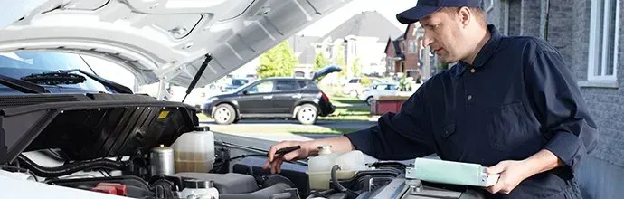 A mechanic in a dark uniform inspects the open hood of a car parked outside a house, holding a diagnostic tablet.