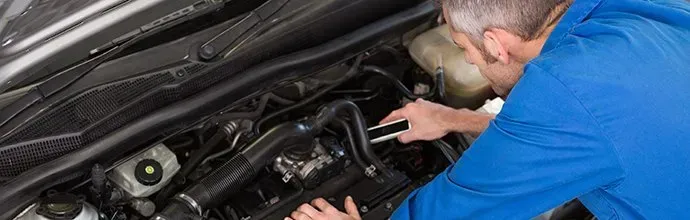 A mechanic in a blue uniform leans over an open car hood to work on the engine.