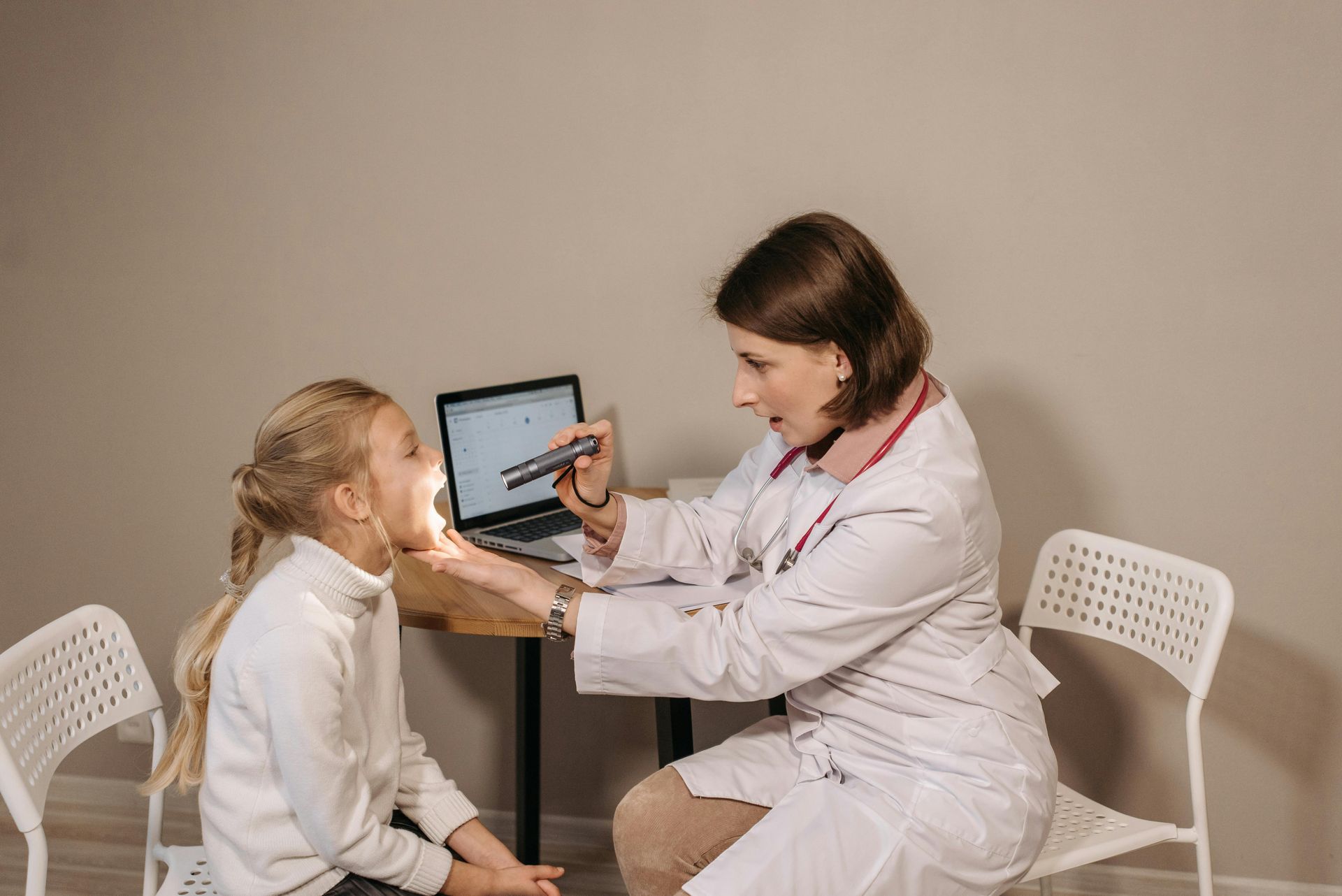 Doctor examining a young girl's throat with a light. The girl sits with mouth open. Office setting.