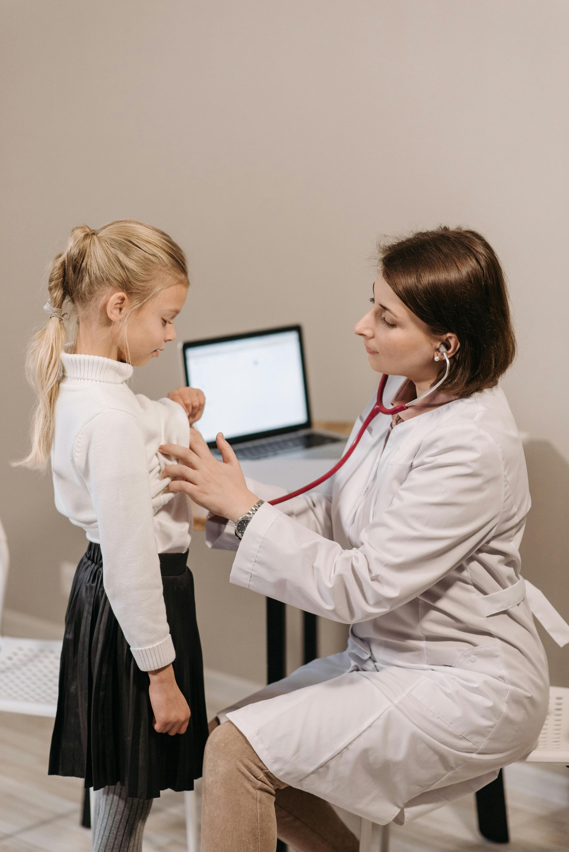 Doctor using stethoscope on child's chest in a neutral-toned room.