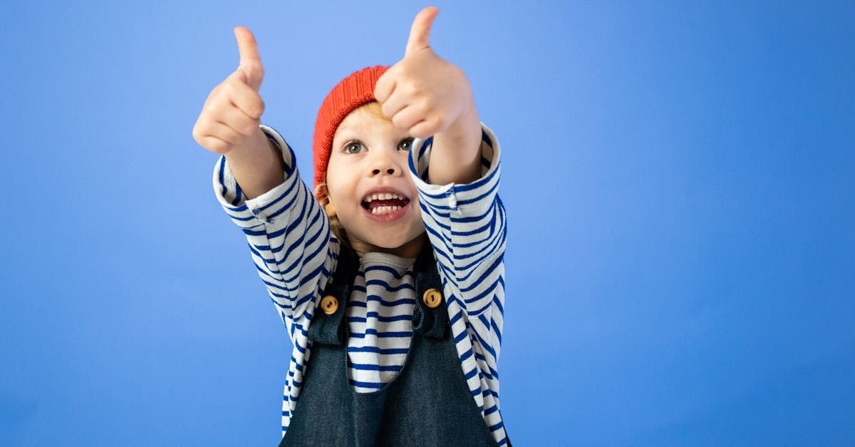Happy child with thumbs up, wearing striped shirt and orange hat against blue backdrop.