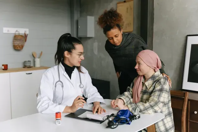 Doctor talking to a young person with cancer, parent nearby; white coat, gray wall, documents on table.
